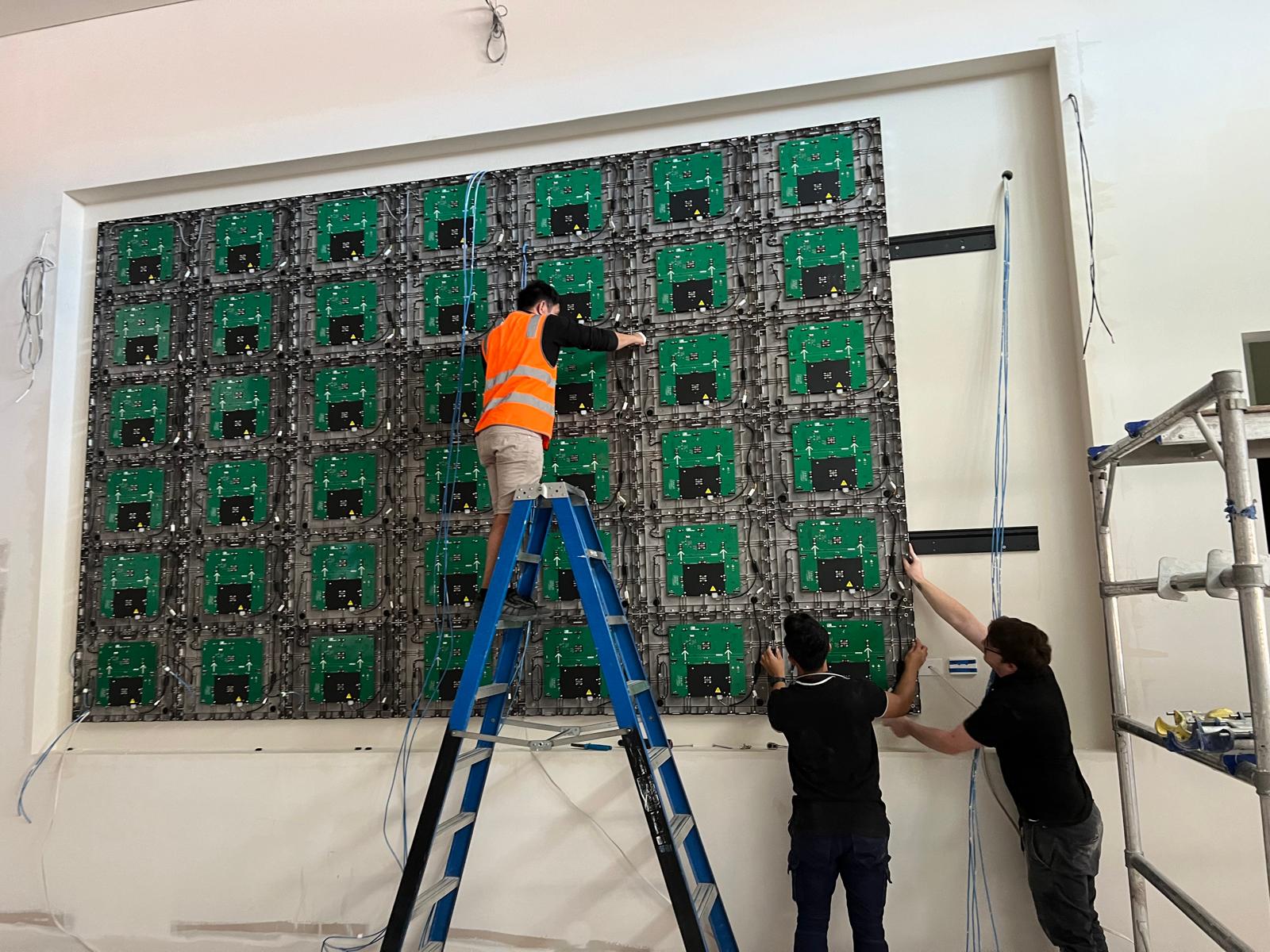 Installers fitting individual LED modules to the sports bar's screen wall, working from a ladder.