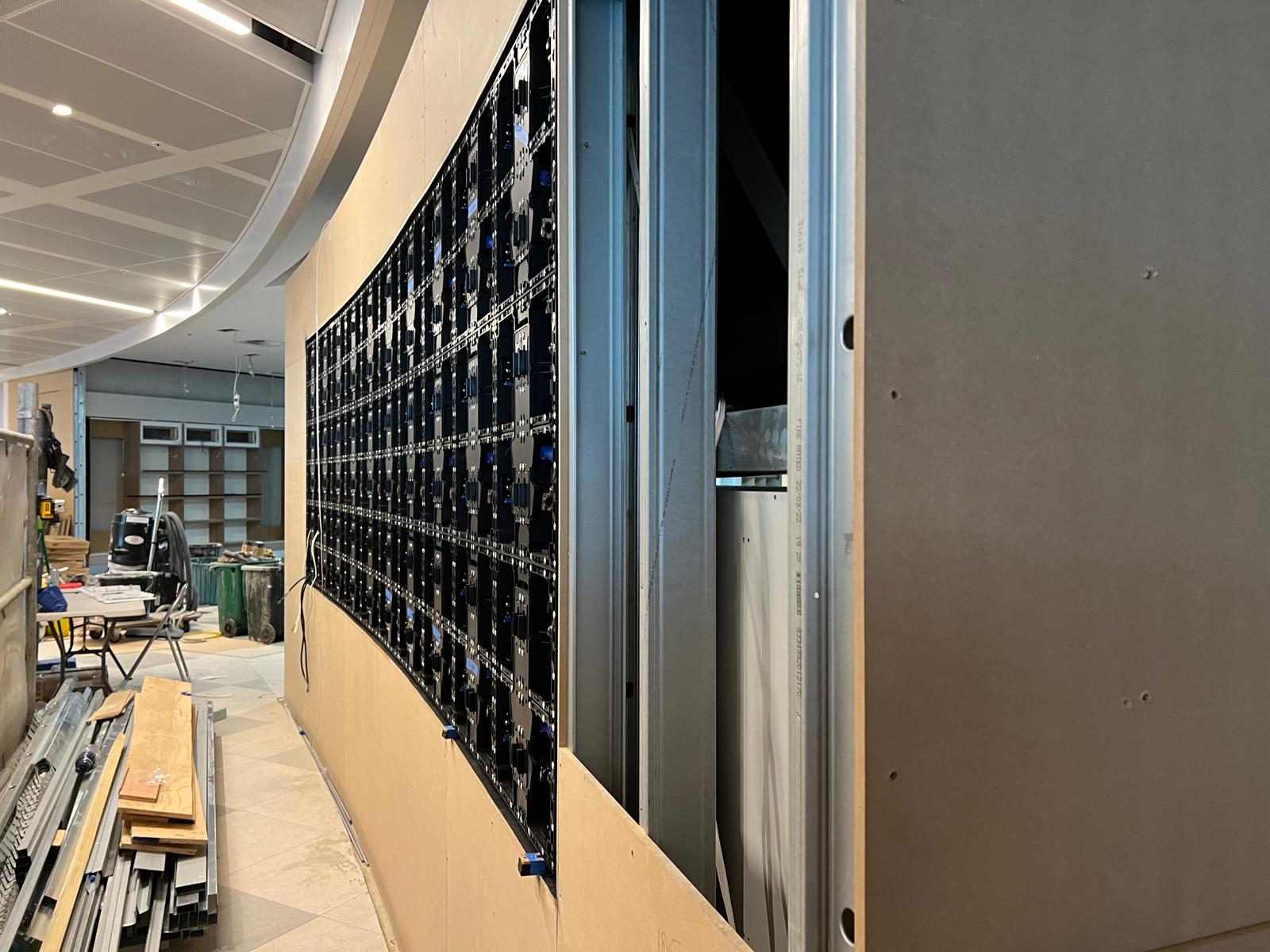 Rear view of a curved LED display panel mounted on its plywood substrate, with the surrounding wall framing still exposed.