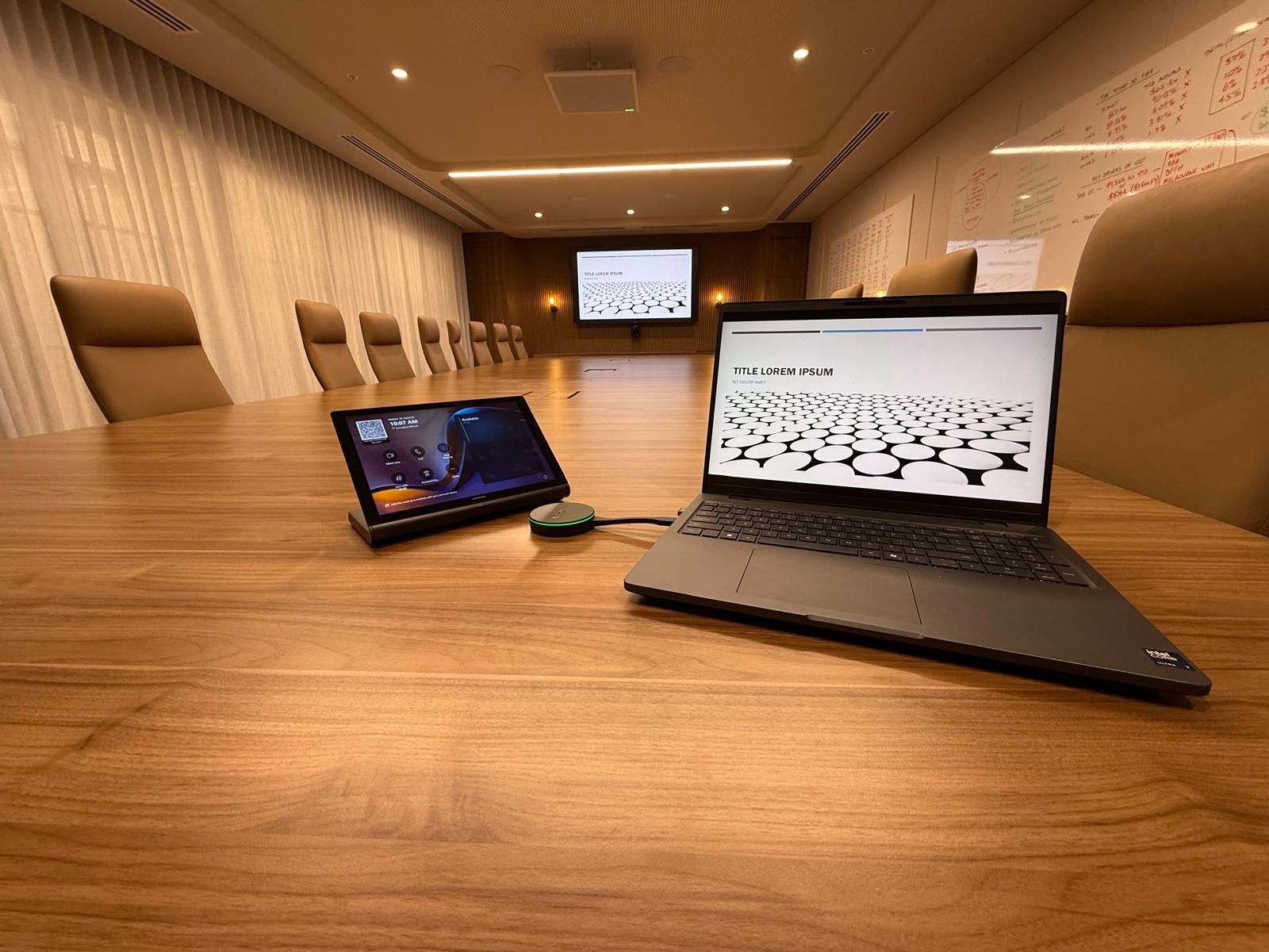 Boardroom table fitted with a video conferencing touch panel and speakerphone, with the presentation display visible at the far end of the room.